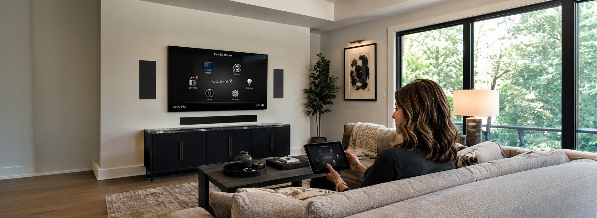 A homeowner uses a tablet to manage lighting, comfort, and security through a Control4 interface displayed on a wall‑mounted TV in a modern living room with large windows and integrated audio.