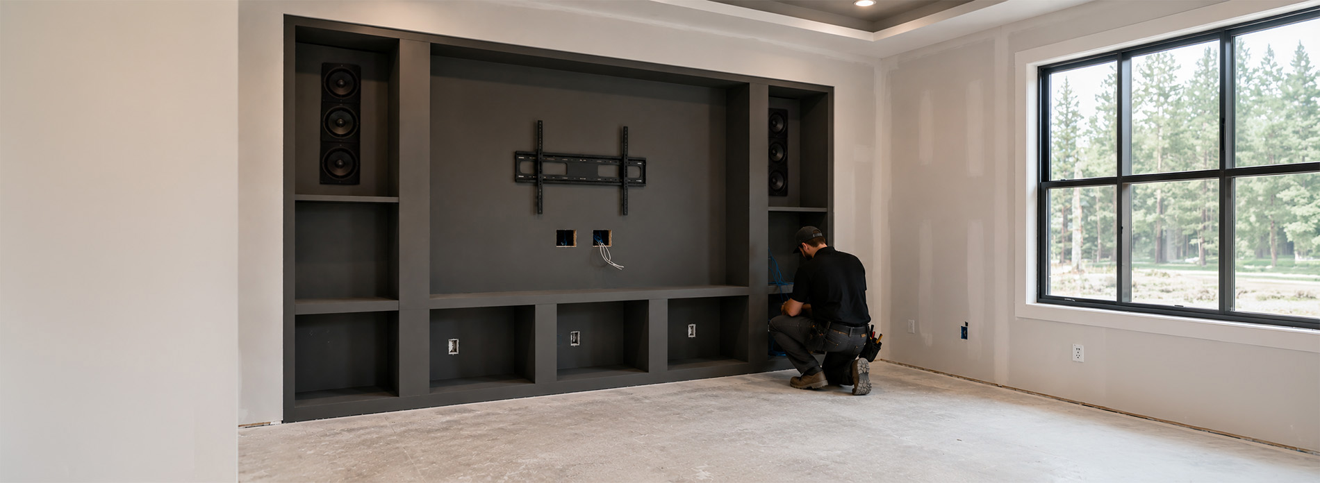 A technician installs wiring and speakers in a built‑in media wall during new‑home construction, with unfinished drywall, recessed lighting, and large windows overlooking pine trees, illustrating smart‑home integration in progress.